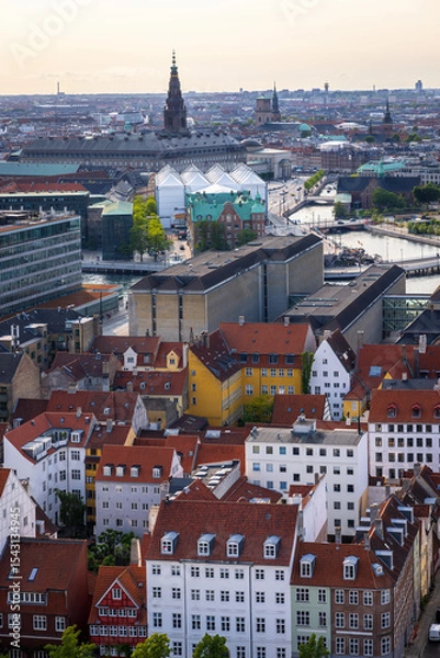 Fototapeta Panoramic view of Copenhagen taken from the top of the Church of Our Saviour, showing a mix of historical rooftops, modern buildings, and city landmarks under a warm evening sky.