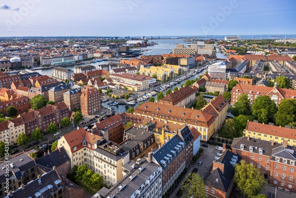 Fototapeta Panoramic view of Copenhagen taken from the top of the Church of Our Saviour, showing a mix of historical rooftops, modern buildings, and city landmarks under a warm evening sky.