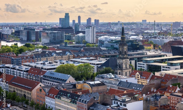 Fototapeta Panoramic view of Copenhagen taken from the top of the Church of Our Saviour, showing a mix of historical rooftops, modern buildings, and city landmarks under a warm evening sky.