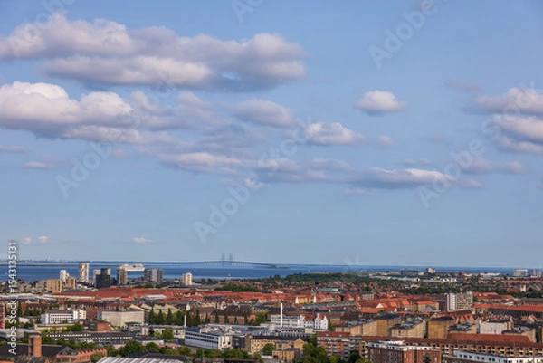 Fototapeta Panoramic view of Copenhagen taken from the top of the Church of Our Saviour, showing a mix of historical rooftops, modern buildings, and city landmarks under a warm evening sky.