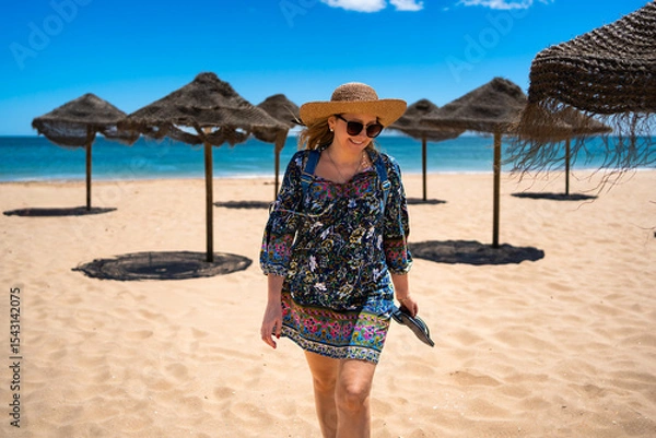 Fototapeta Beautiful happy middle aged woman tourist in straw hat walking on sandy beach full of beach umbrellas in summertime. Front view. Meia Praia beach in Lagos, Portugal