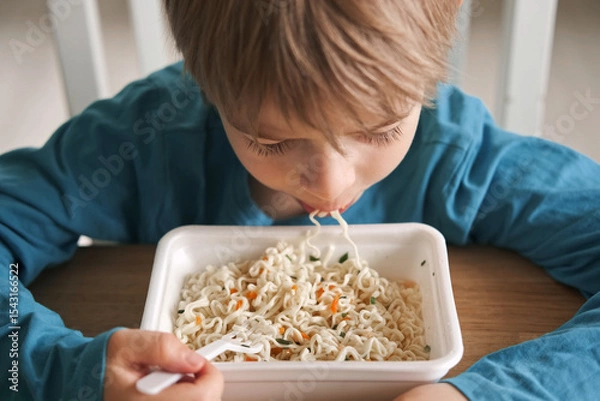 Fototapeta 6 years old boy eats instant noodle from box. Child is eating spaghetti. Lunch time. A good appetite. Tasty children food. Close-up portrait. Carbs dinner. Fast food. Kid eating by himself with fork.