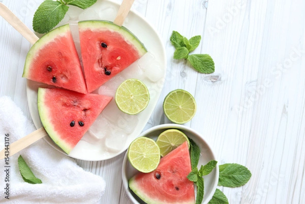 Fototapeta Ripe watermelon pieces on sticks like ice cream. Watermelon, lime and mint. Summer fruits background. Flat lay. Watermelon slices in white plate top view, wooden table and lime halves. Summer food