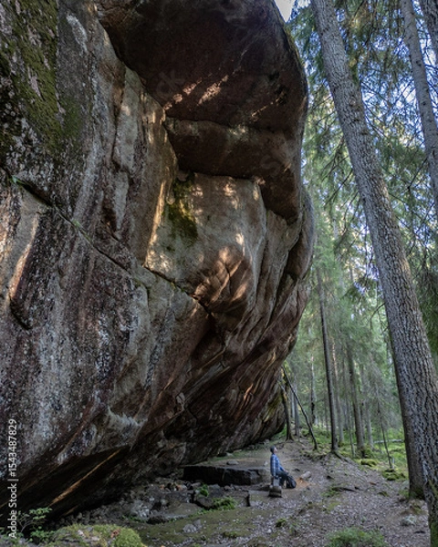 Fototapeta A man standing at the base of a massive rock formation known as Pirunkirkko (Devil’s Church) in Paistjärvi, Finland. This stunning geological site lies deep in the forest, with textured stone surfaces