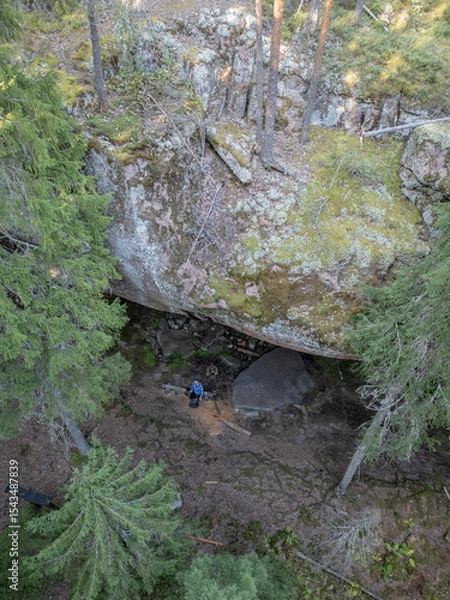 Fototapeta Aerial view of a person standing beneath the dramatic rock overhang of Pirunkirkko (Devil’s Church) in Paistjärvi, Finland. The moss-covered cliffs and surrounding pine forest.
