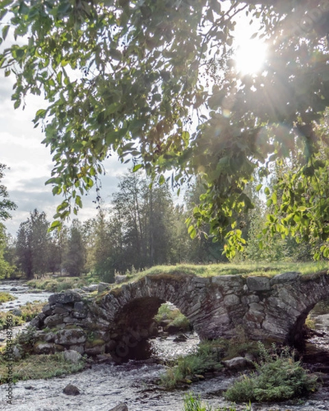 Fototapeta A rustic stone arch bridge spans a gently flowing river in the Finnish countryside, with sunlight filtering through green tree leaves. The peaceful rural scene evokes timeless natural beauty.