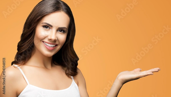 Fototapeta Photo of smiling woman in white casual wear holding hand with flat palm, giving, advertising some product showing mock up copy space free area. Isolated brown background. Brunette girl.