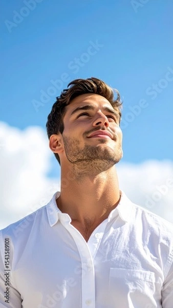 Fototapeta Skyward Gaze: A handsome man, bathed in natural light, gazes skyward with hopeful and positive aspirations under the vast canvas of a bright blue sky, embodying hope and aspiration.