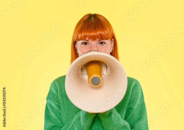 Obraz Woman with bright red hair holds a megaphone in front of a yellow background while wearing a green sweater, conveying a message or call to action