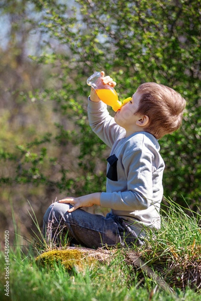 Obraz A little boy sits on the grass and greedily drinks orange juice