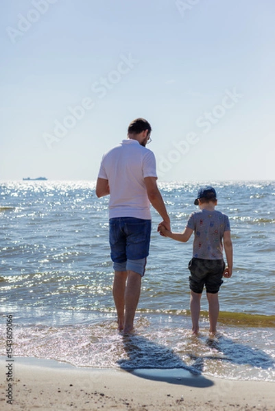 Obraz A father and son are wading in the water by the Baltic Sea, holding hands, and looking at the water.