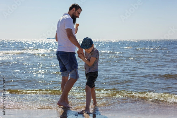 Obraz A bearded father and his little son are wading in the water and eating ice cream at the seaside
