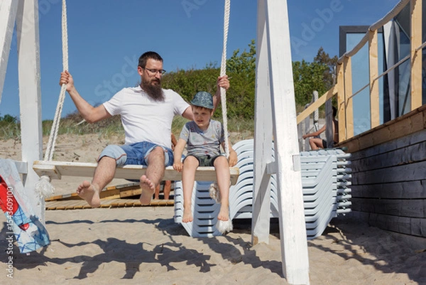 Obraz A bearded father with his son swings on a beach swings