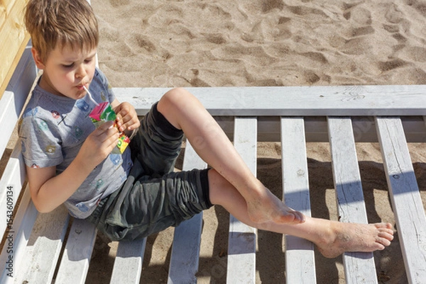Obraz A cute little boy sits on a wooden beach chair and drinks juice from a small packet