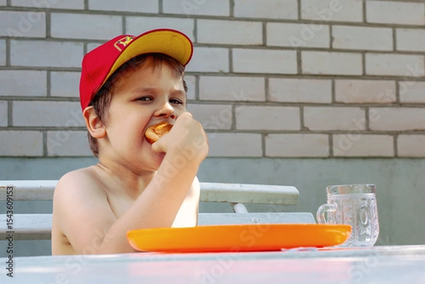 Obraz A little boy in a red cap is eating a big delicious bun