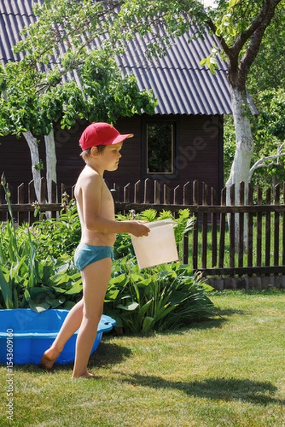Obraz A little boy with a red hat and a white bucket in his hands is getting ready to cool off in the pool in his grandmother's garden on a hot summer day