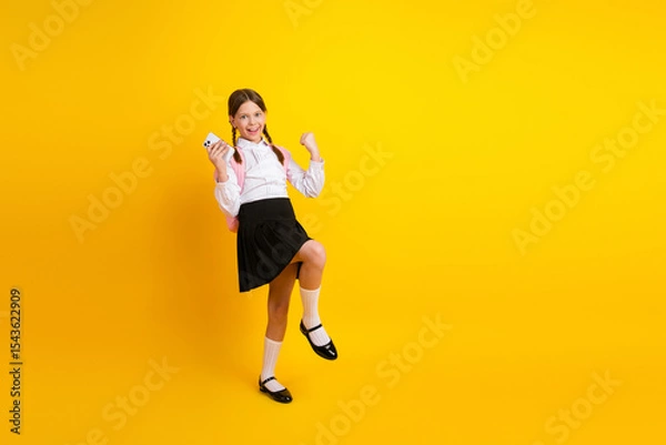 Fototapeta Cheerful schoolgirl holding a phone and posing on a yellow background in stylish uniform with a backpack