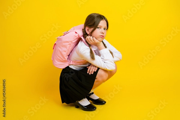 Obraz Schoolgirl sitting against bright yellow background wearing a school uniform and pink backpack, looking thoughtful and stylish