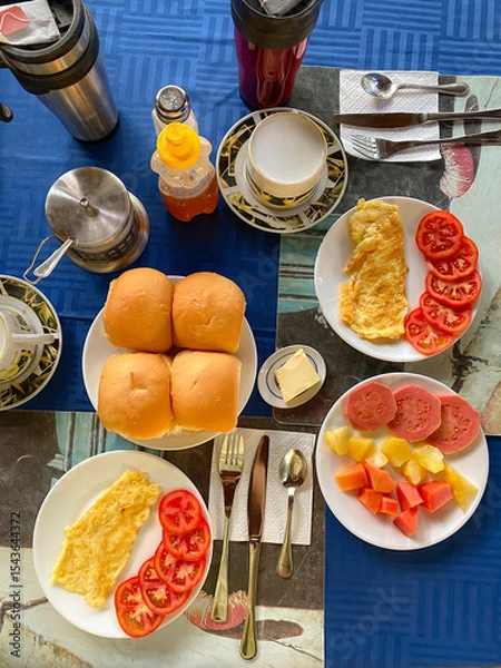 Fototapeta Breakfast in hotel. Vertical, top view of healthy breakfast spread at homestay or guesthouse. Eggs, fresh fruits, bread tea and juice. Cozy, rustic setting, dining, or food concept. Trinidad, Cuba.