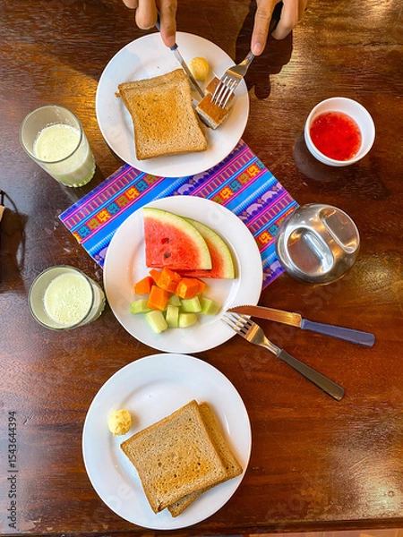 Fototapeta Healthy breakfast in hotel concept. Simple meal of fresh fruits, toasted bread, fruit juice. Person cutting food on plate. Top down angle flat lay of breakfast setting, wooden table. La Paz, Bolivia.