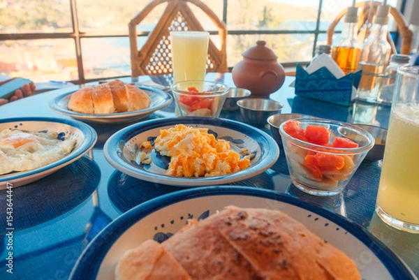 Fototapeta Healthy hotel breakfast with eggs, fruits, bread and juice on a sunlit table with window view. Rustic setting, travel accommodation or hospitality concept. Copacabana, Bolivia. Cozy hotel stay.