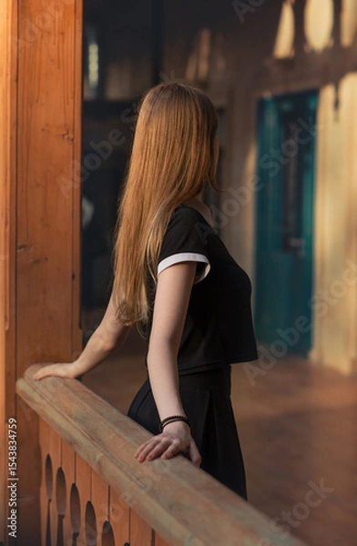 Fototapeta Young woman stands on a wooden balcony, gazing toward a door bathed in warm light. A symbolic moment of dreams, inner desires, and stepping into the future