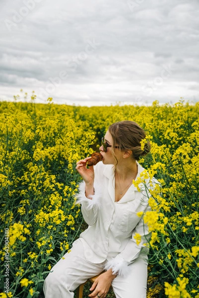 Fototapeta Young woman enjoys snack in rapeseed field under cloudy sky during late afternoon