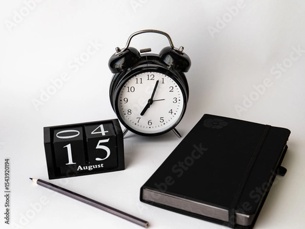 Obraz Still life depicting a classic black alarm clock, a wooden cubic calendar showing "August 15th", a black diary or notebook and a pencil, all placed on a light (white) background.