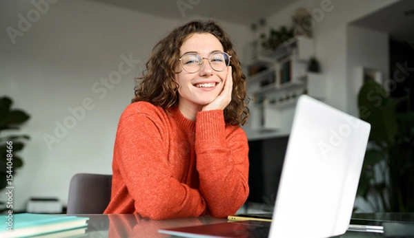 Obraz Portrait of happy young woman or student wearing glasses sitting at desk at home office with laptop computer looking at camera advertising online learning, remote work, business webinars.