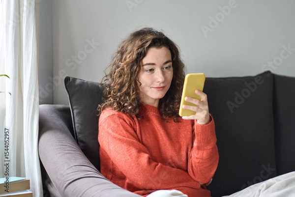 Obraz Pretty young woman sitting on sofa using cell phone, holding smartphone, looking at cellphone doing online shopping choosing fashion clothes in mobile apps, checking social media commercials.