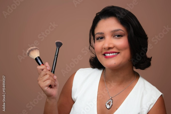 Fototapeta Friendly woman in white tank top smiling and holding make-up brushes. Suitable for beauty, cosmetics, makeup and embellishment themes. Horizontal studio portrait with copy space on beige background.
