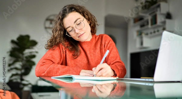 Obraz Busy young woman using laptop sitting at desk writing notes while watching webinar, studying online, learning web classes or having virtual call meeting remote working from home.