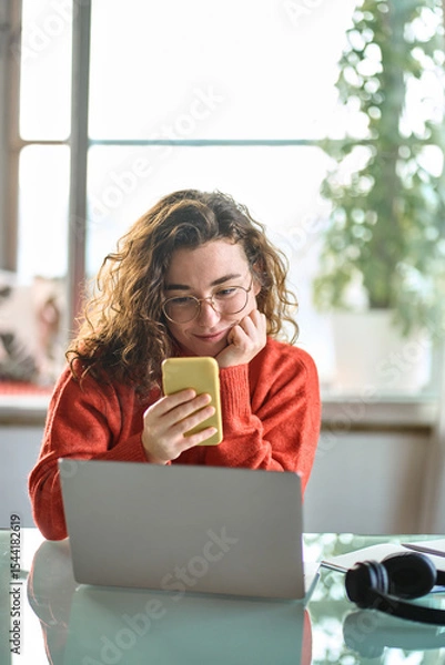 Obraz Vertical photo of young smiling pretty woman holding smartphone using cellphone modern technology, looking at mobile, checking cell phone apps, watching social media sitting at table at home.