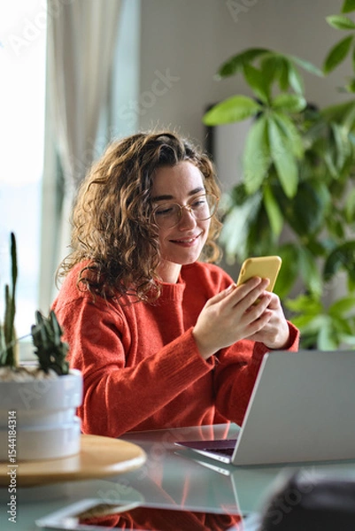 Obraz Happy young lady using smartphone social media apps at home. Smiling woman holding cellphone modern ai applications, looking at mobile, checking cell phone, texting chatting sitting at table, vertical