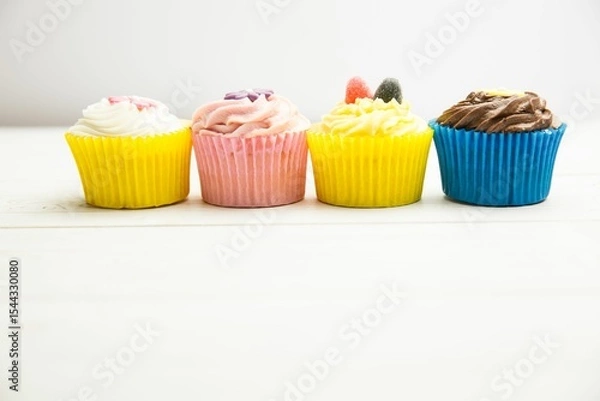 Fototapeta Cupcakes are being shown in flat design on white wooden tabletop with colorful liners and frosting