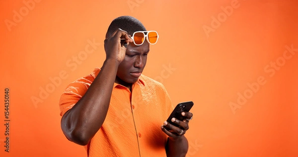 Obraz Black man, phone and confused in studio with chat, thinking and reading post on orange background. African person, smartphone and doubt with fake news, gossip and scroll choice on social network
