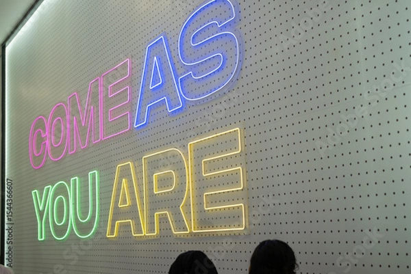 Fototapeta Side angle photo of a neon wall sign displaying the words "COME AS YOU ARE" in colorful lights on a perforated background inside a store.