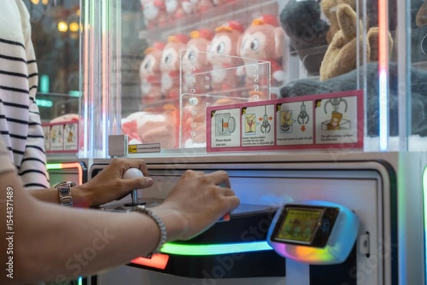 Fototapeta Hands operating a joystick and button on a claw machine at an arcade, with colorful plush toys and instruction stickers in the background.
