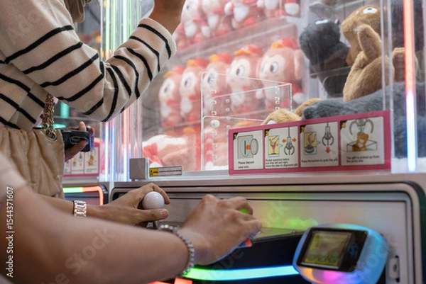 Fototapeta Two people using a joystick and buttons to play a colorful claw machine, surrounded by plush toys. The game area is bright and vibrant, capturing a fun and competitive arcade moment.