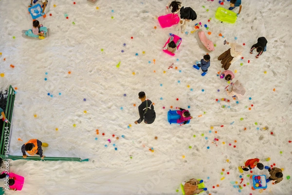 Fototapeta Top view of children playing on artificial snow with colorful toys and sleds in an indoor playground area, creating a vibrant and joyful winter-themed atmosphere.
