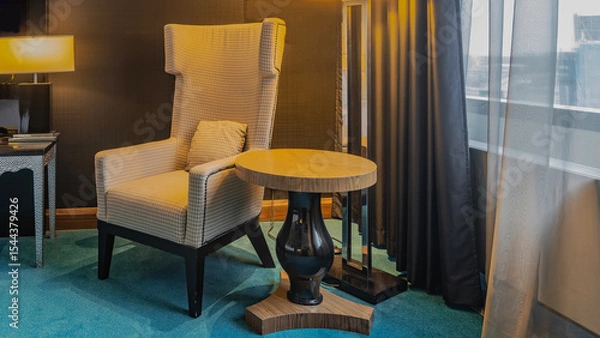 Fototapeta The interior of the living room. A soft armchair with a cushion in the corner. A round coffee table on one leg next to it. The lamp is lit. There are tulle curtains on the window. Blue carpet 