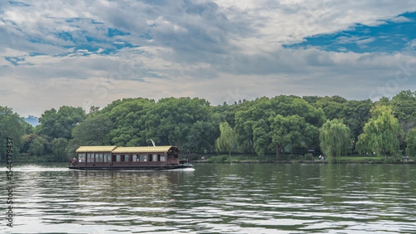 Fototapeta A beautiful Chinese wooden tourist boat with a tiled roof floats on a calm lake. Glare, ripples on the water. Green vegetation on the shore. Blue sky, clouds. China. Hangzhou. West Lake Xi Hu
