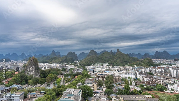 Fototapeta Panorama of a modern city. Lots of buildings, green trees. There is a pavilion on top of a high sheer cliff. Silhouettes of mountains on the horizon. Clouds, sky. China.Guilin.View from the Fubo Hill 