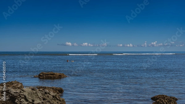 Fototapeta A man swims on his back in the blue ocean. The person in sunglasses is smiling. Ripples on calm water. Foam waves in the distance. Boulders in the foreground. Azure sky, clouds. Copy space. Madagascar