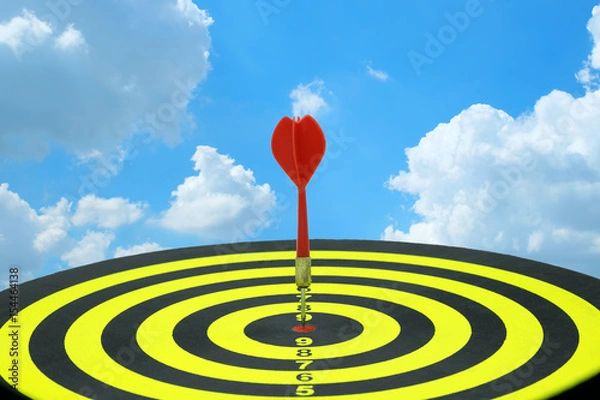 Obraz Red dart arrow hitting in the target center of dartboard with blue sky and clouds background for conceptual of vision, target, finish in business