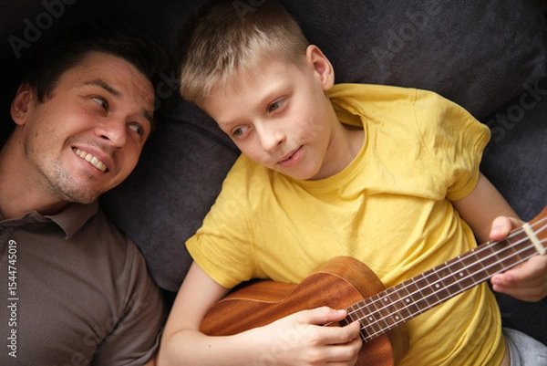 Fototapeta Happy father watching smiling son playing ukulele at home, relaxing on sofa together