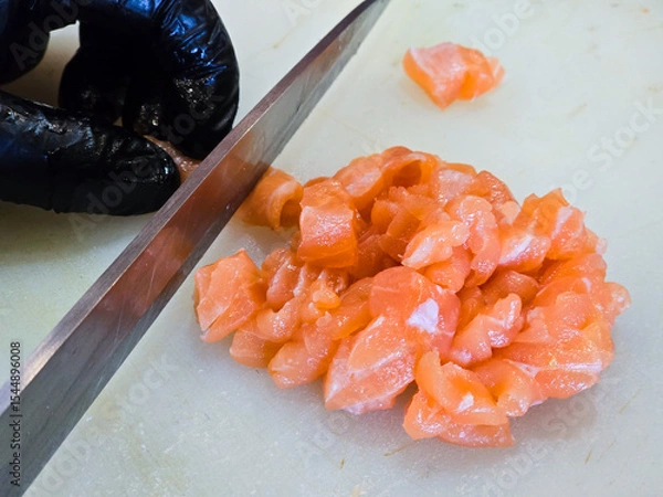 Fototapeta Chef slicing raw salmon into small cubes on a white cutting board, wearing black gloves for hygiene, preparing ingredients for sashimi or poke dishes.