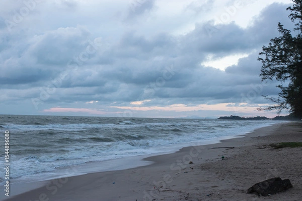 Fototapeta A quiet beach with strong waves under a cloudy sky in the early evening, with pine trees lining the coast and soft light peeking through the clouds