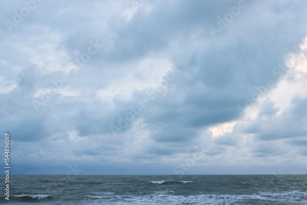 Fototapeta A quiet beach with strong waves under a cloudy sky in the early evening, with pine trees lining the coast and soft light peeking through the clouds