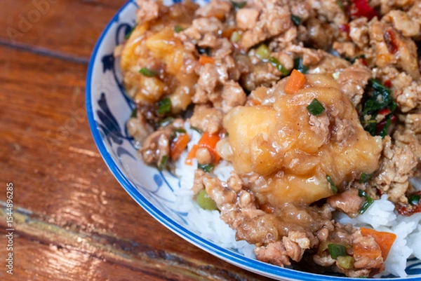 Fototapeta A Thai rice dish topped with stir-fried minced pork, vegetables, and battered fried egg, served in a patterned ceramic bowl on a wooden table.